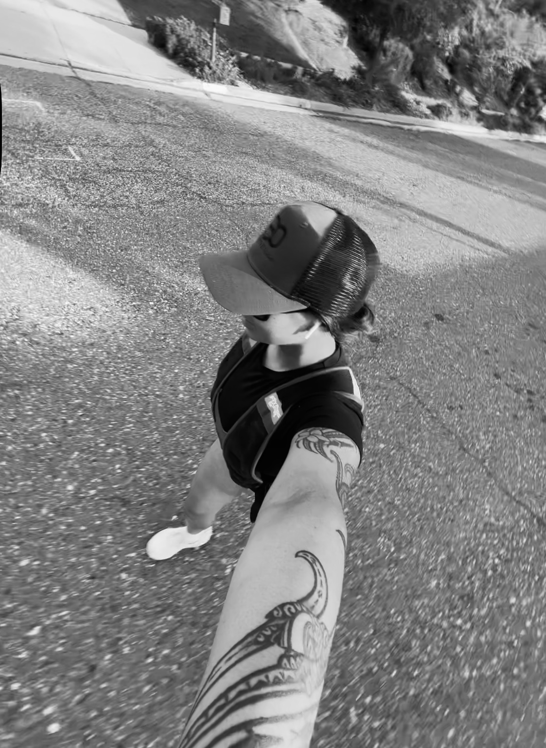 Black and white photo of a person walking outdoors on a paved road, wearing a baseball cap, athletic outfit, and a weighted vest. The photo is taken from a high angle, showing the person’s tattooed arm extended forward as they hold the camera.