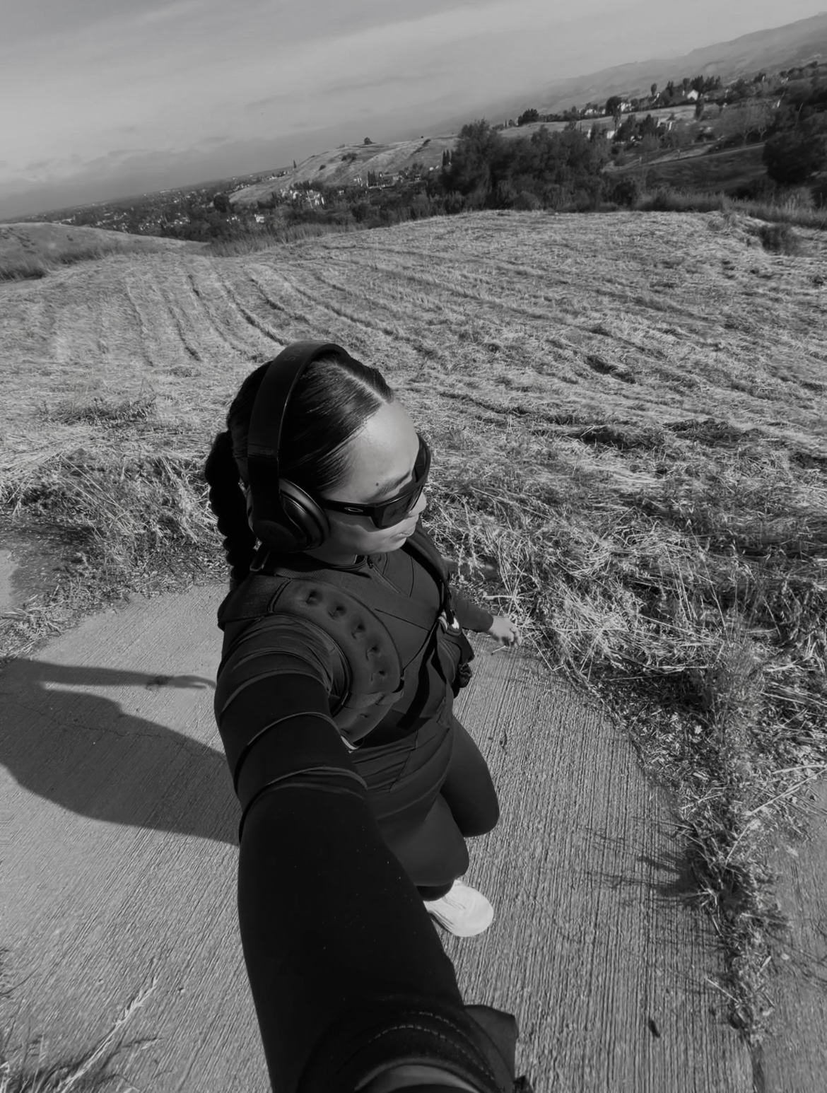 A woman wearing headphones, sunglasses, and athletic gear with a weighted vest takes a selfie while walking on a paved path through a rural landscape with harvested fields and distant hills under a partly cloudy sky.