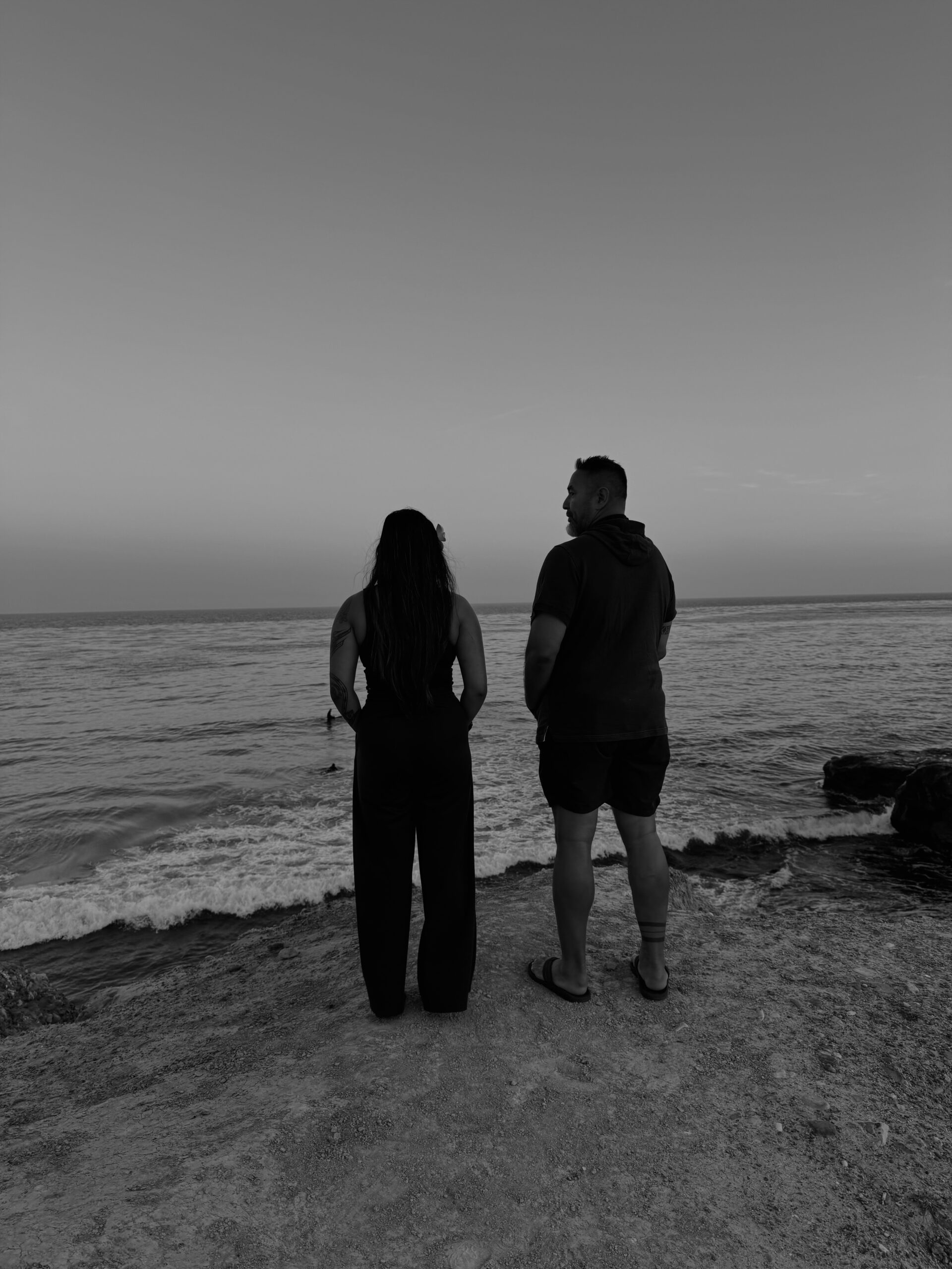 A black-and-white photo of a man and woman standing on a rocky shoreline, gazing out at the ocean during sunset. Waves gently crash below as surfers are visible in the water. The sky is calm and expansive, evoking a sense of peace and reflection.