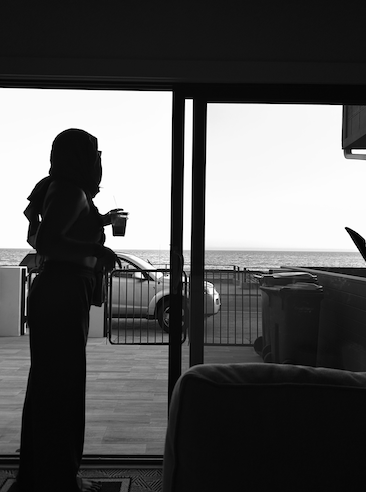 Silhouette of a woman holding a drink, looking out at the ocean through a glass door—symbolizing reflection and spiritual insight.