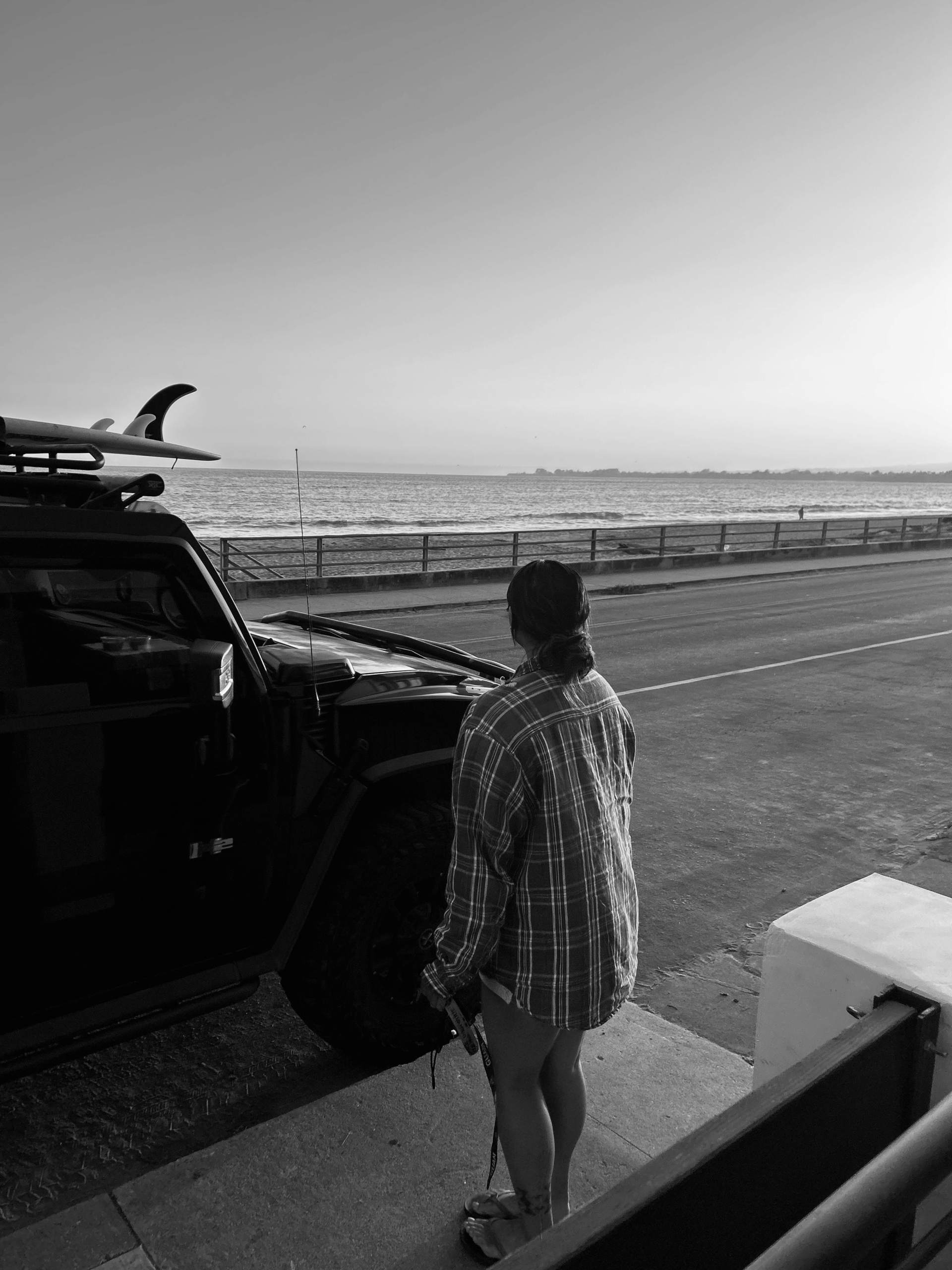 Woman in a plaid shirt standing by a black SUV with surfboards on top, looking out at the ocean during sunrise or sunset.