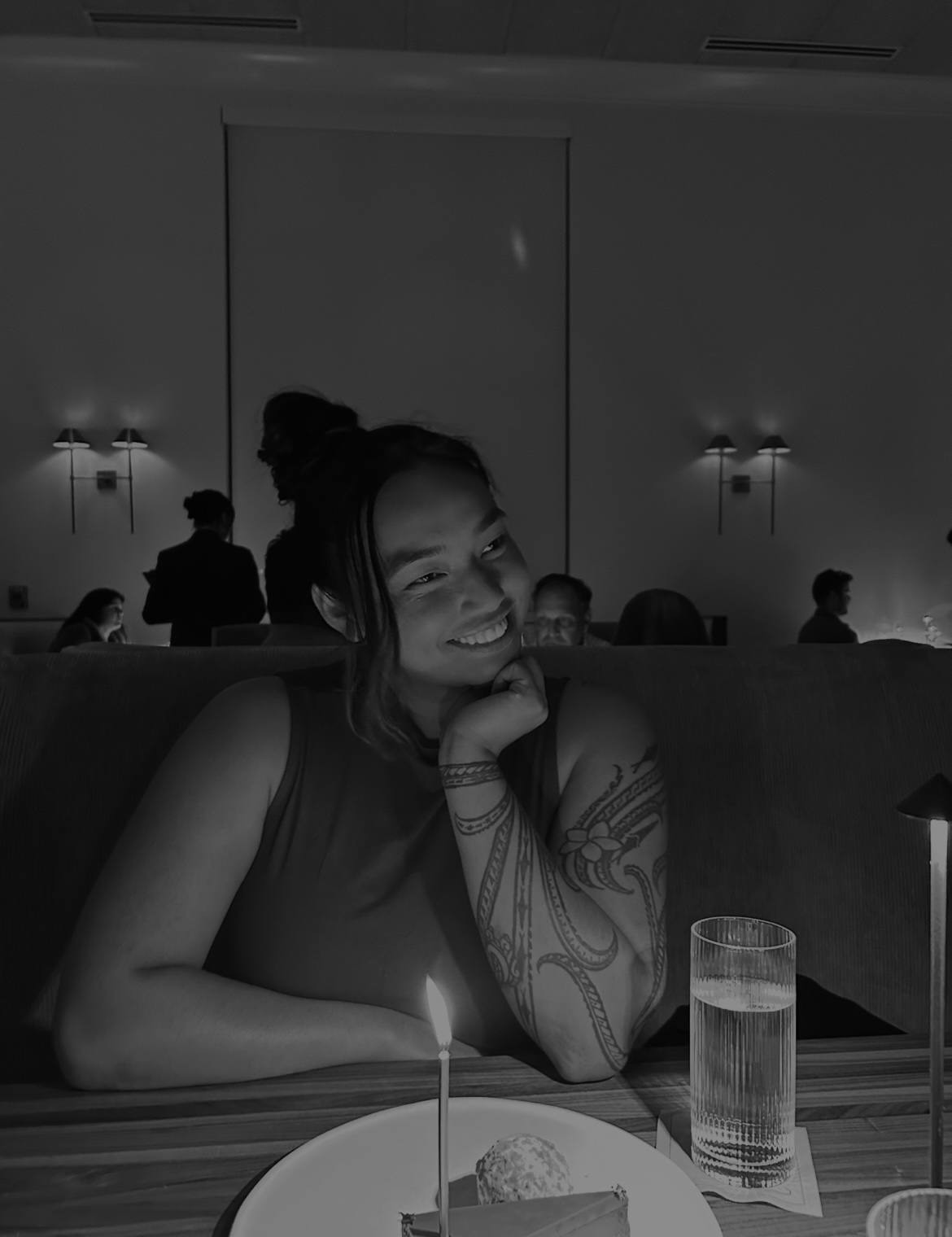 Black-and-white photo of a woman smiling thoughtfully while seated at a restaurant booth, resting her chin on her hand. A small lit candle stands in front of a dessert on the table, with a glass of water nearby. Soft lighting and other diners appear blurred in the background, creating a calm, intimate atmosphere.
