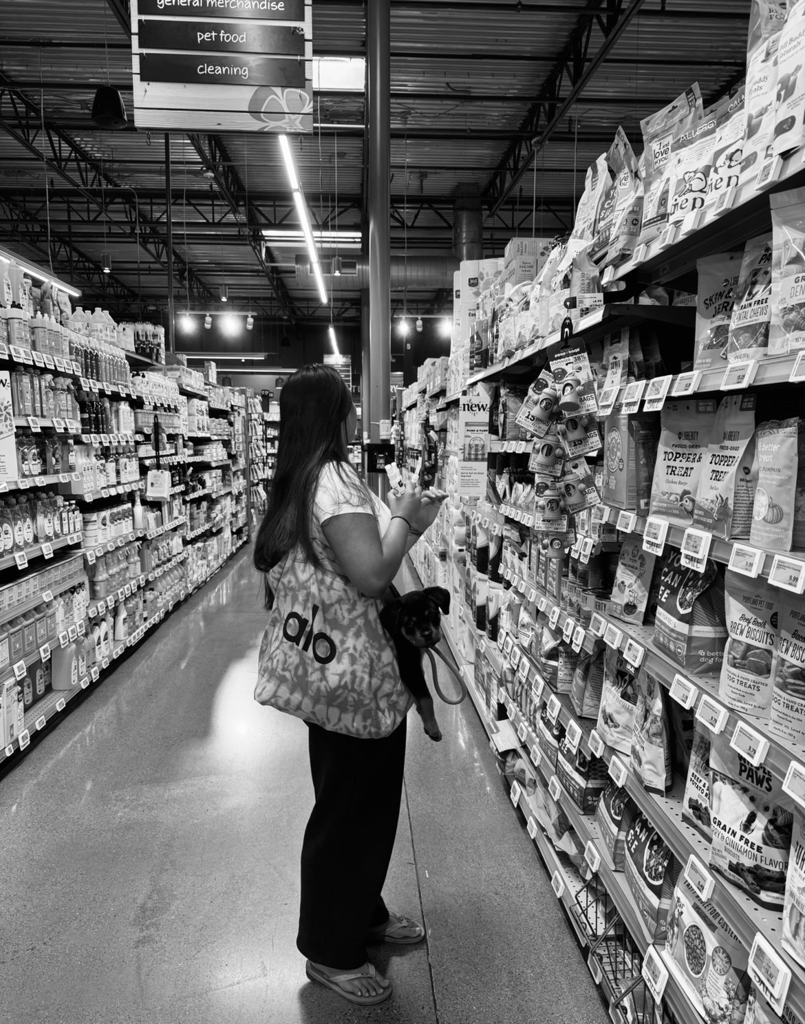 A shopper browses pet food options while holding her dog in a well-stocked pet store aisle.